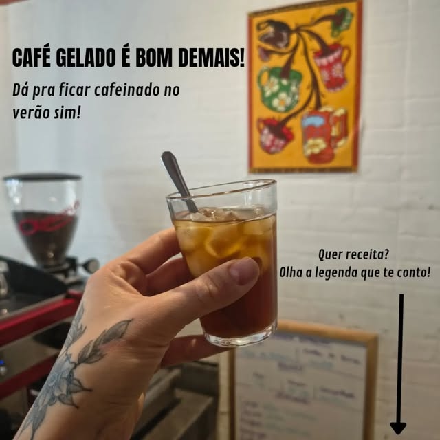Copo de café gelado com gelo e colher, segurado por uma mão, diante de um quadro colorido de xícaras