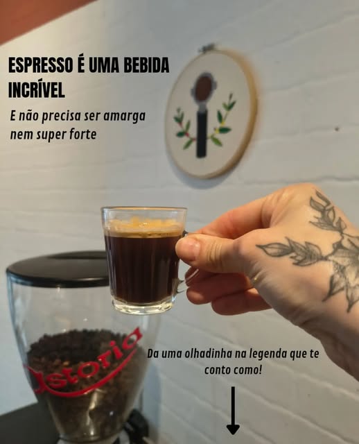 Mão segurando um espresso em copo de vidro, ao lado de um moedor com grãos e um bordado de porta-filtro na parede branca