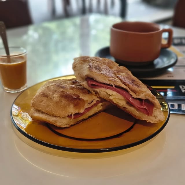 Sanduíche de mortadela com queijo cortado ao meio sobre prato âmbar, com xícara cor de terracota ao fundo