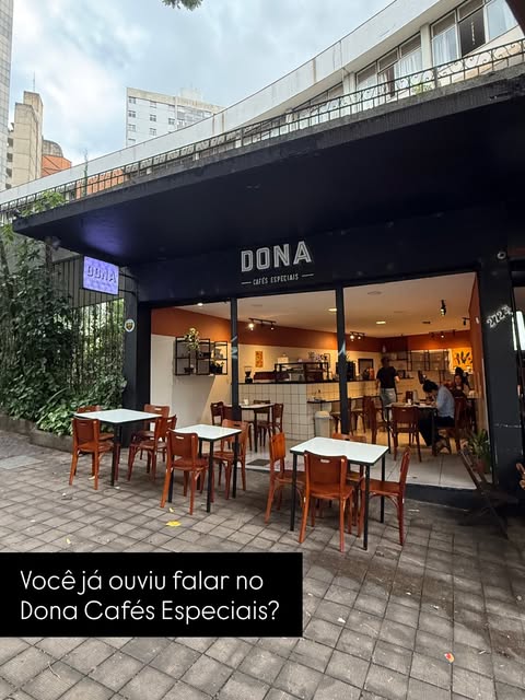 Fachada da Dona com mesas de madeira na calçada e o salão aceso ao fundo